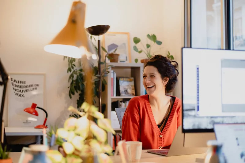A team member laughs at their desk, with "Bringing new voices to the fore" displayed on the wall.