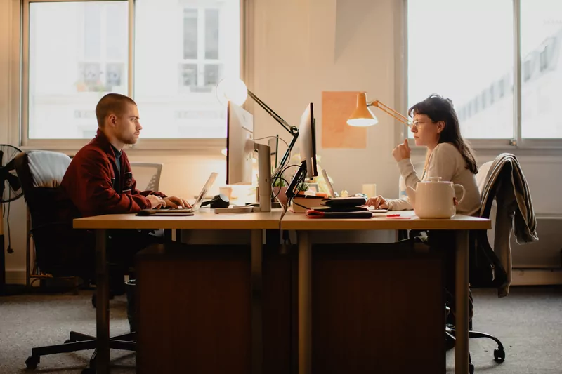 Two people work facing each other at their desks, with windows in a row.